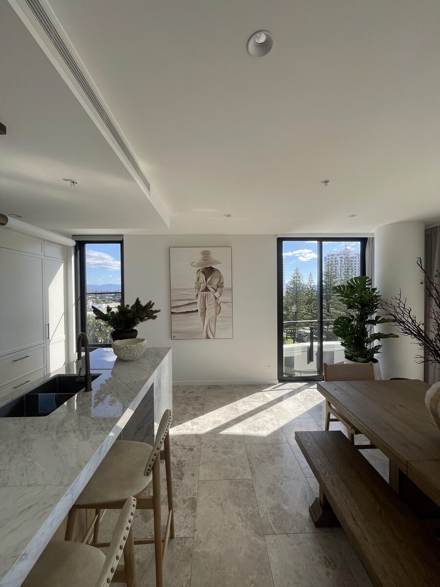 Tall framed coastal figure artwork installed between two windows in an open-plan apartment kitchen with marble island.