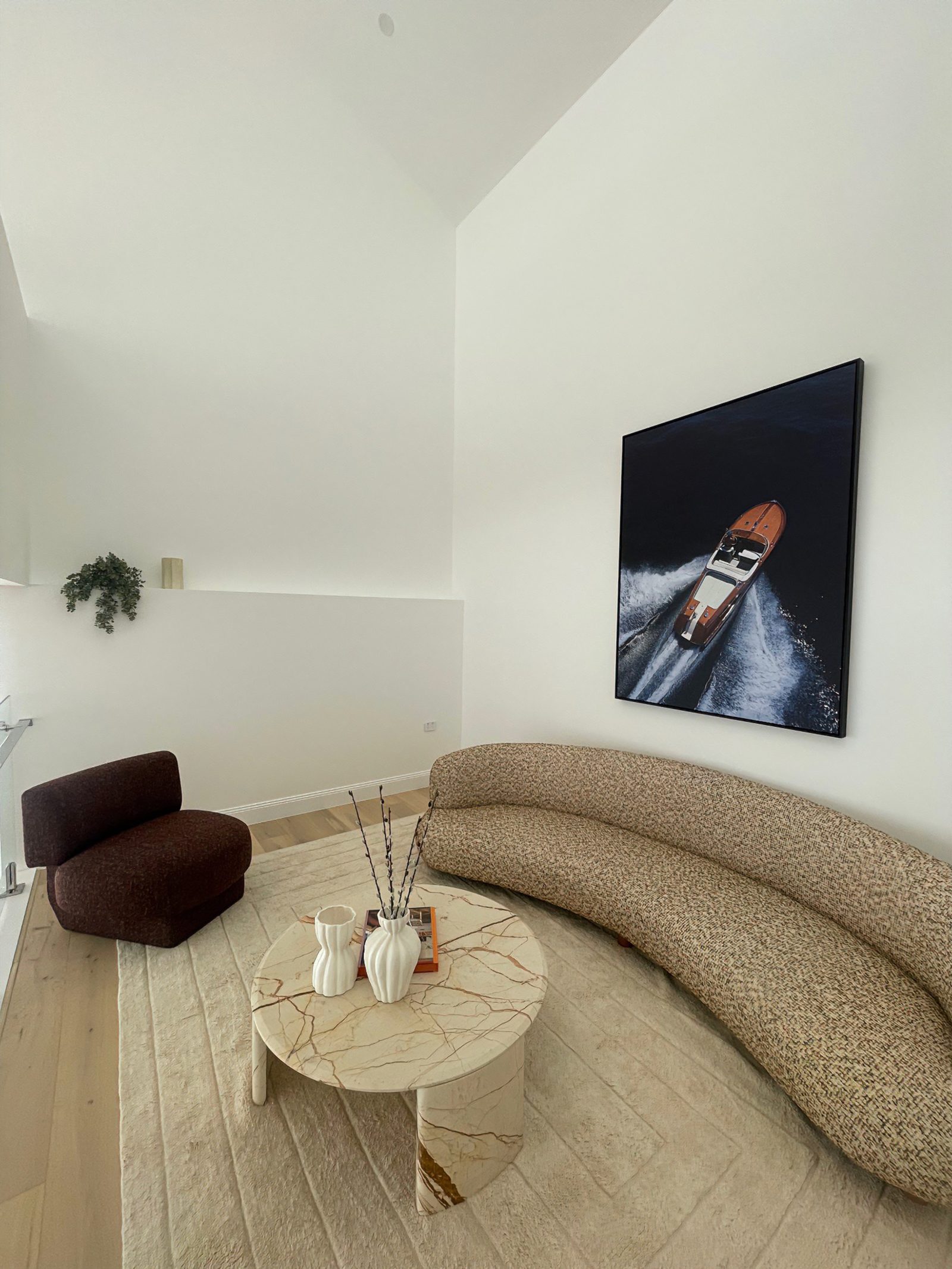 Large framed aerial photograph of a speedboat hung on a tall void wall, above a curved boucle sofa and marble coffee table.