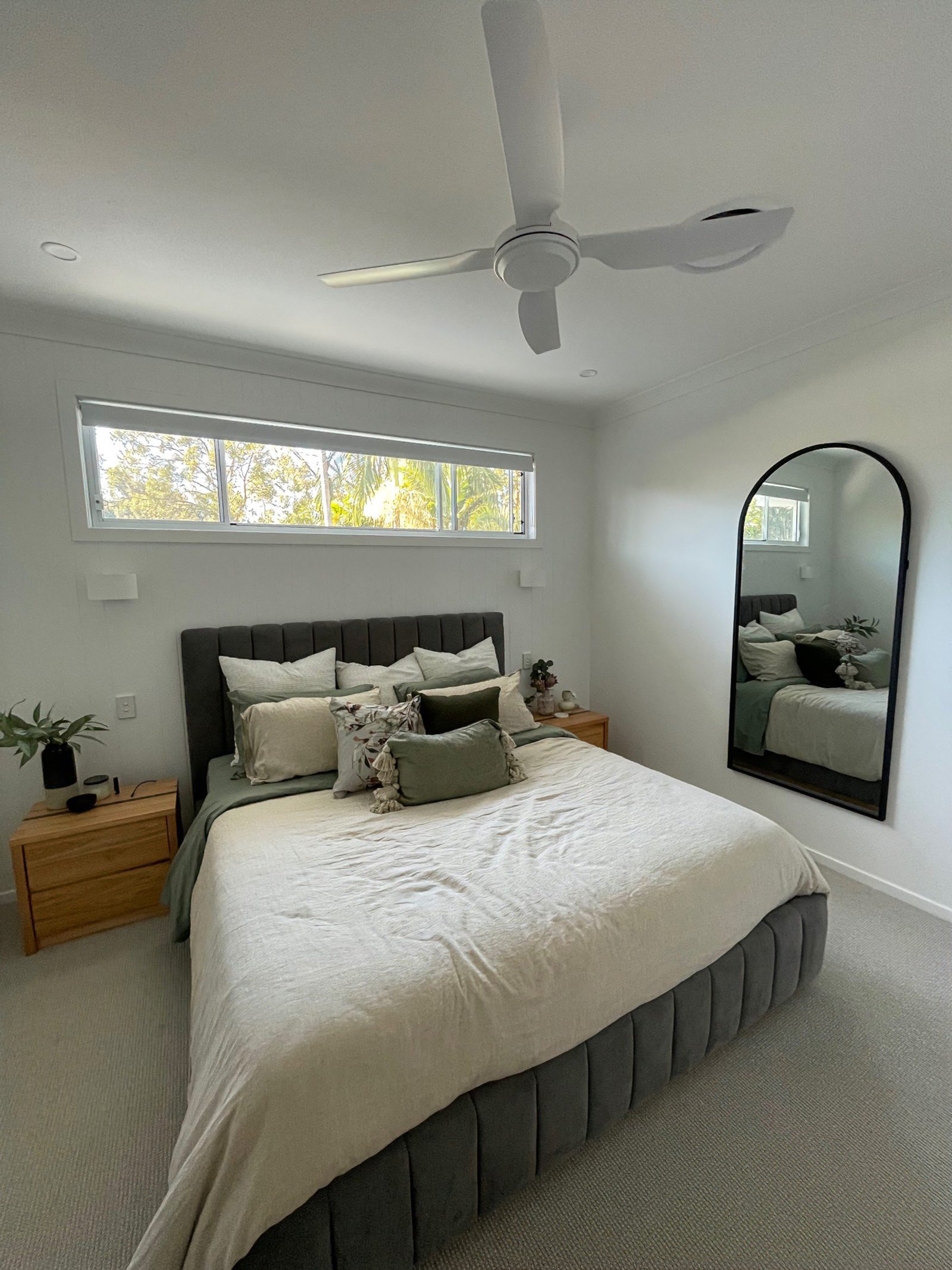 Charcoal channel-tufted upholstered bedhead fixed level on a bedroom wall, with a tall arched mirror mounted opposite reflecting the bed.
