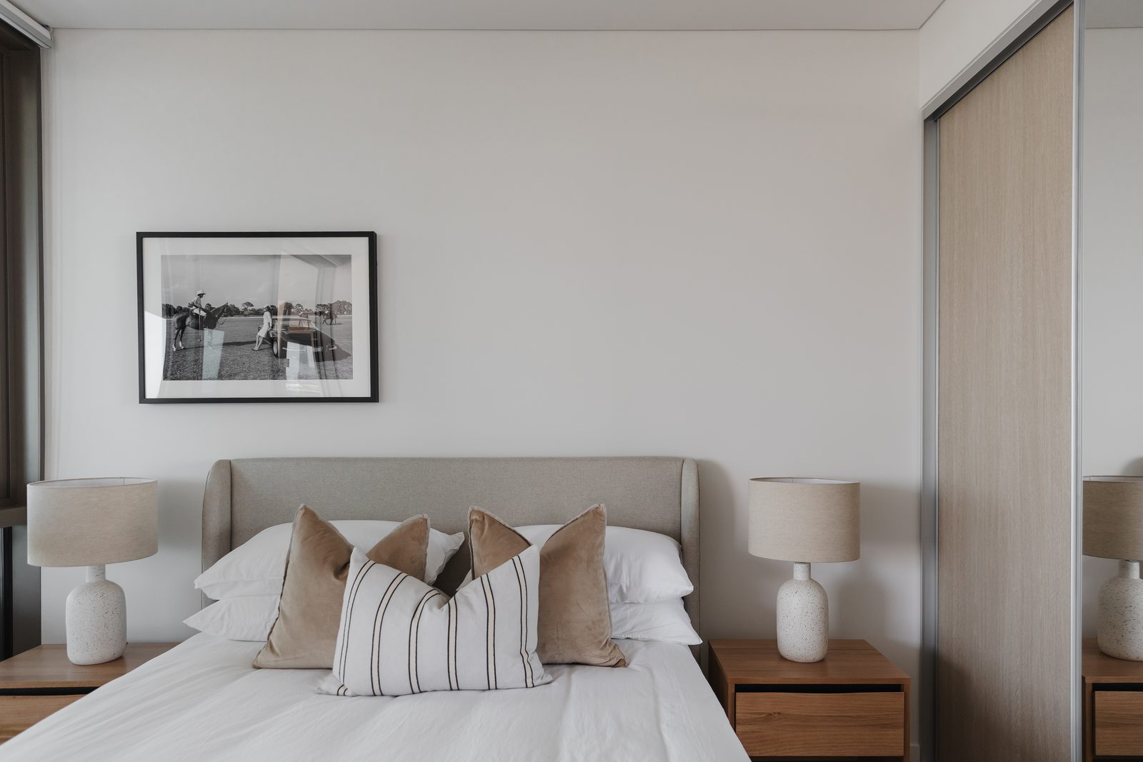 Beige linen bedhead fixed level on the wall with a framed black-and-white polo photograph centred above and ceramic textured lamps either side.