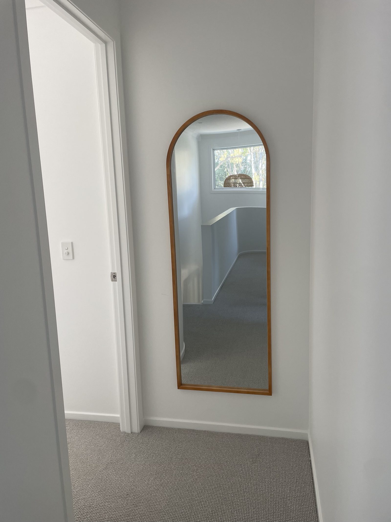 Tall arched timber-framed mirror mounted in a hallway nook, reflecting the upstairs landing and a pendant light.