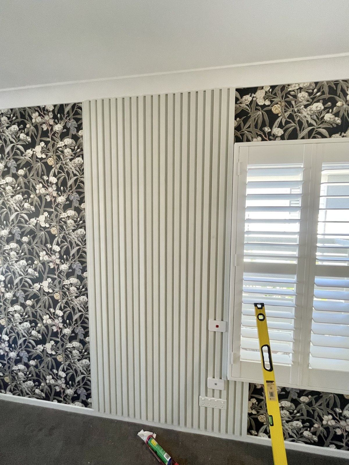 A bedroom slat wall mid-install, with wallpaper on one half and white slats set true on the other.