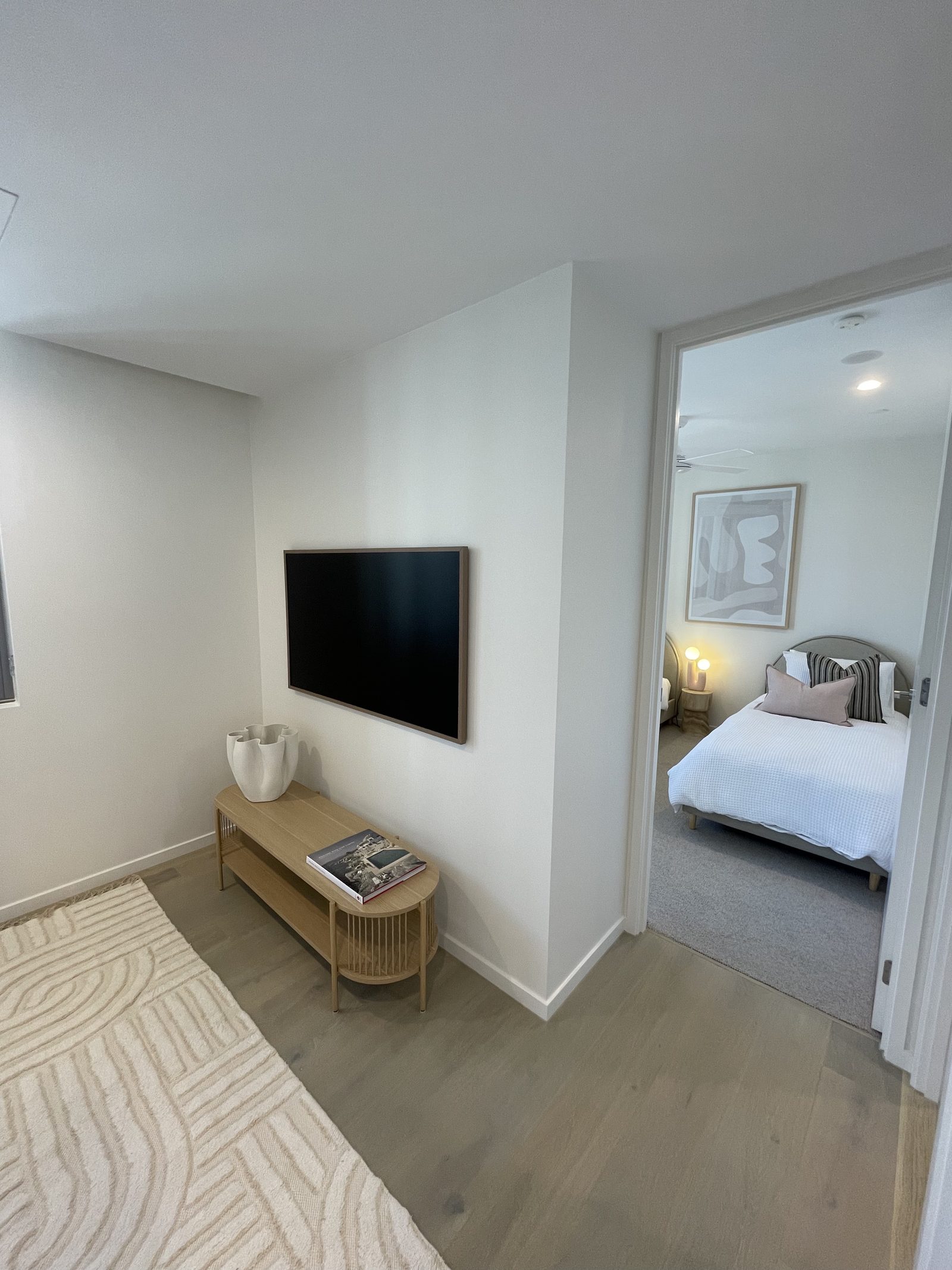 Wall-mounted TV set in a small alcove above a timber console, with a framed abstract artwork visible above a bedhead in the room beyond.