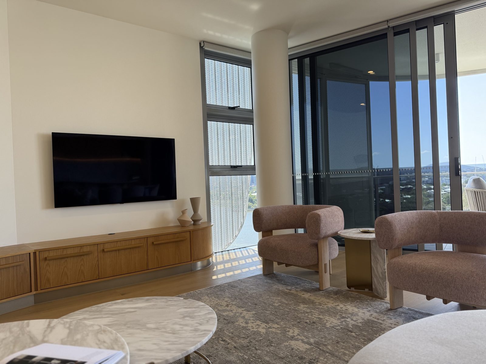 Wall-mounted TV in a modern high-rise apartment with a timber media console below and curved boucle chairs framing the room.