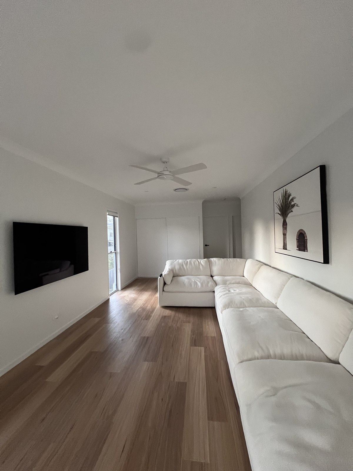 Wall-mounted TV opposite framed palm-tree artwork above a long white sectional, contemporary lounge.