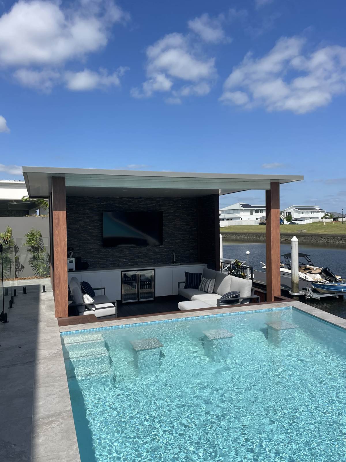 Wide poolside view of a wall-mounted TV in a canal-front cabana lounge, stacked-stone feature wall and timber columns either side.