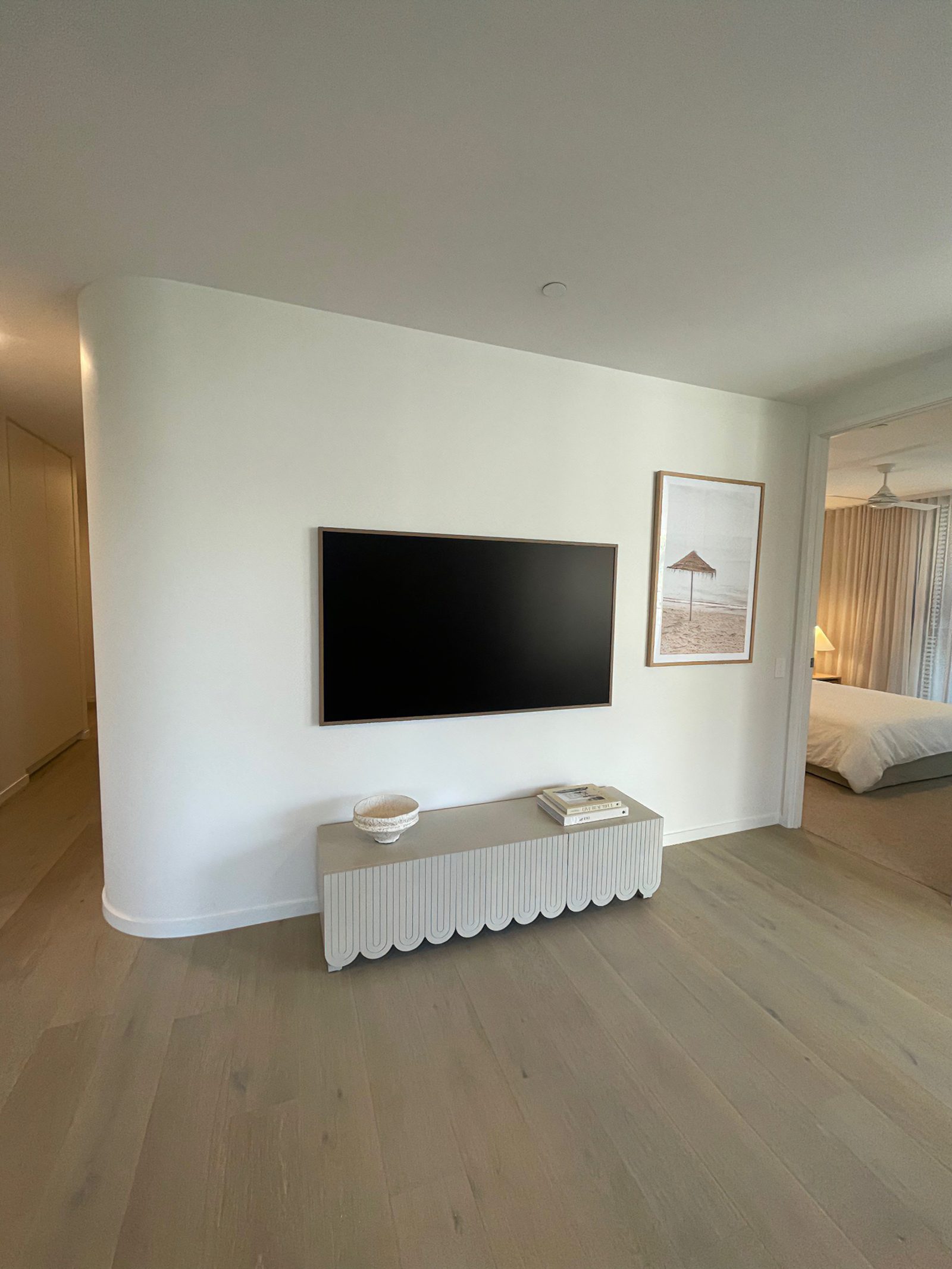 Wall-mounted TV on a curved white feature wall, set above a scalloped console with a framed beach umbrella print beside it and a bedroom visible beyond.