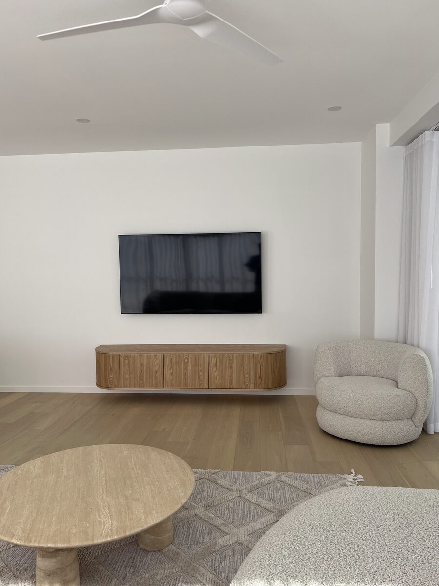 Floating timber console wall-fixed beneath a flush TV install in a coastal living room. Cliff Hanger Installations.