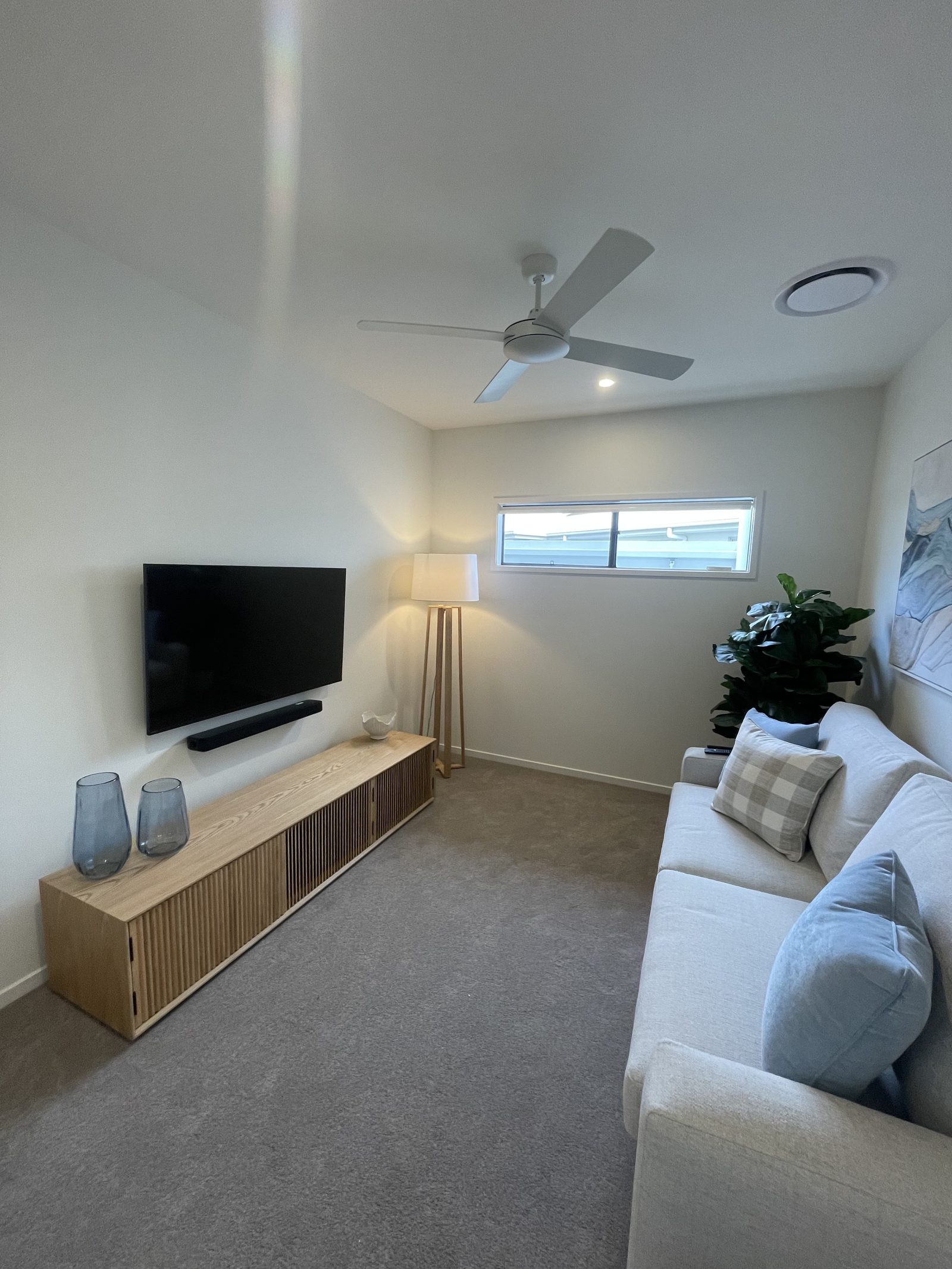 Wall-mounted TV with soundbar in a carpeted media room, set above a timber slat console with framed coastal artwork beside.