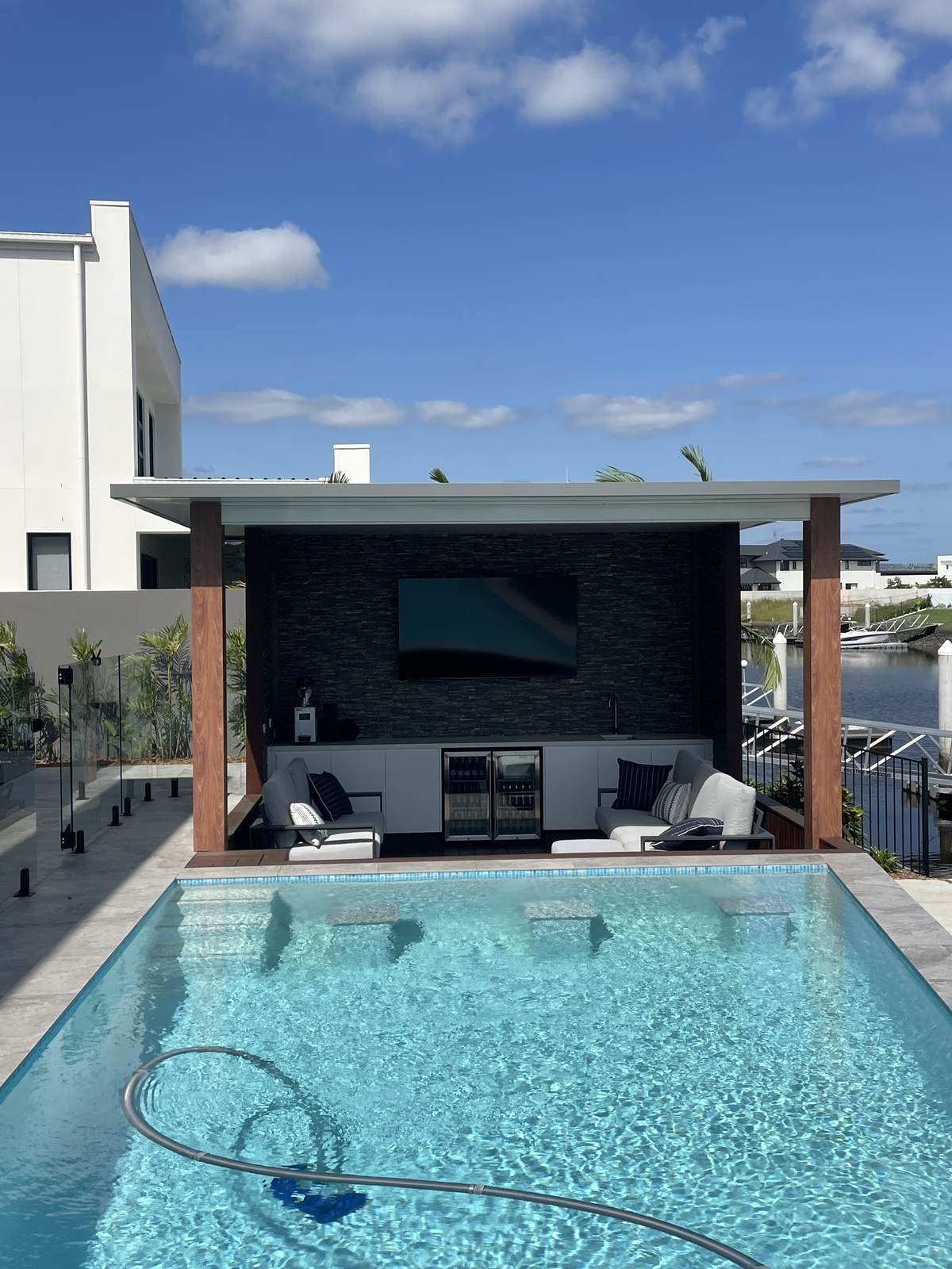 Wall-mounted TV in a poolside cabana on a Gold Coast canal, set on a stacked-stone feature wall with a private mooring behind.