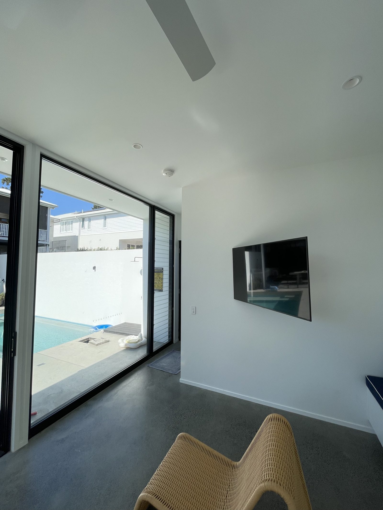 Wall-mounted TV in a cabana opening onto a pool through floor-to-ceiling glass, with rattan lounger in the foreground.