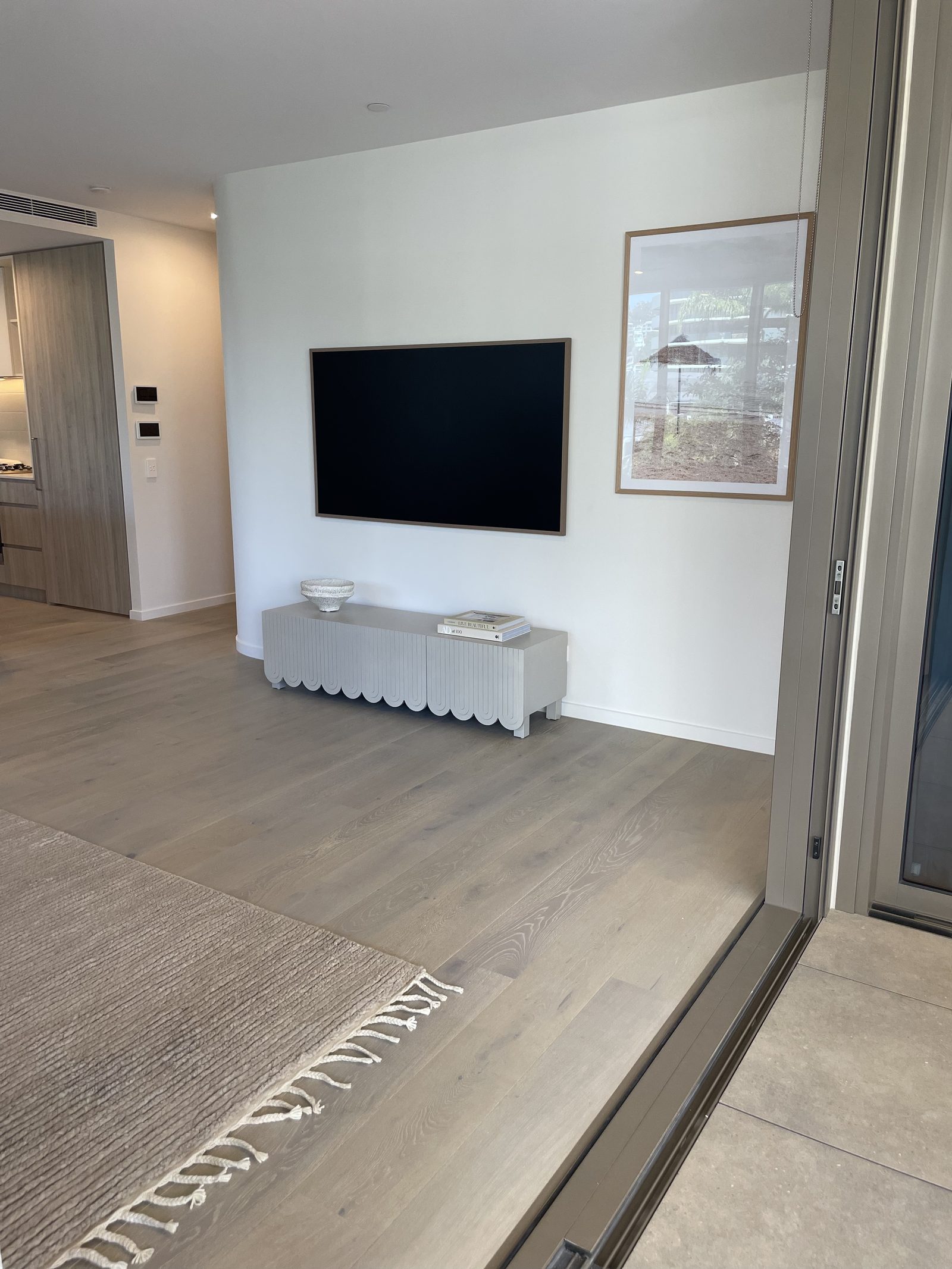 Wall-mounted TV centred above a scalloped white console in a light-filled apartment, with a framed beach umbrella print beside it.