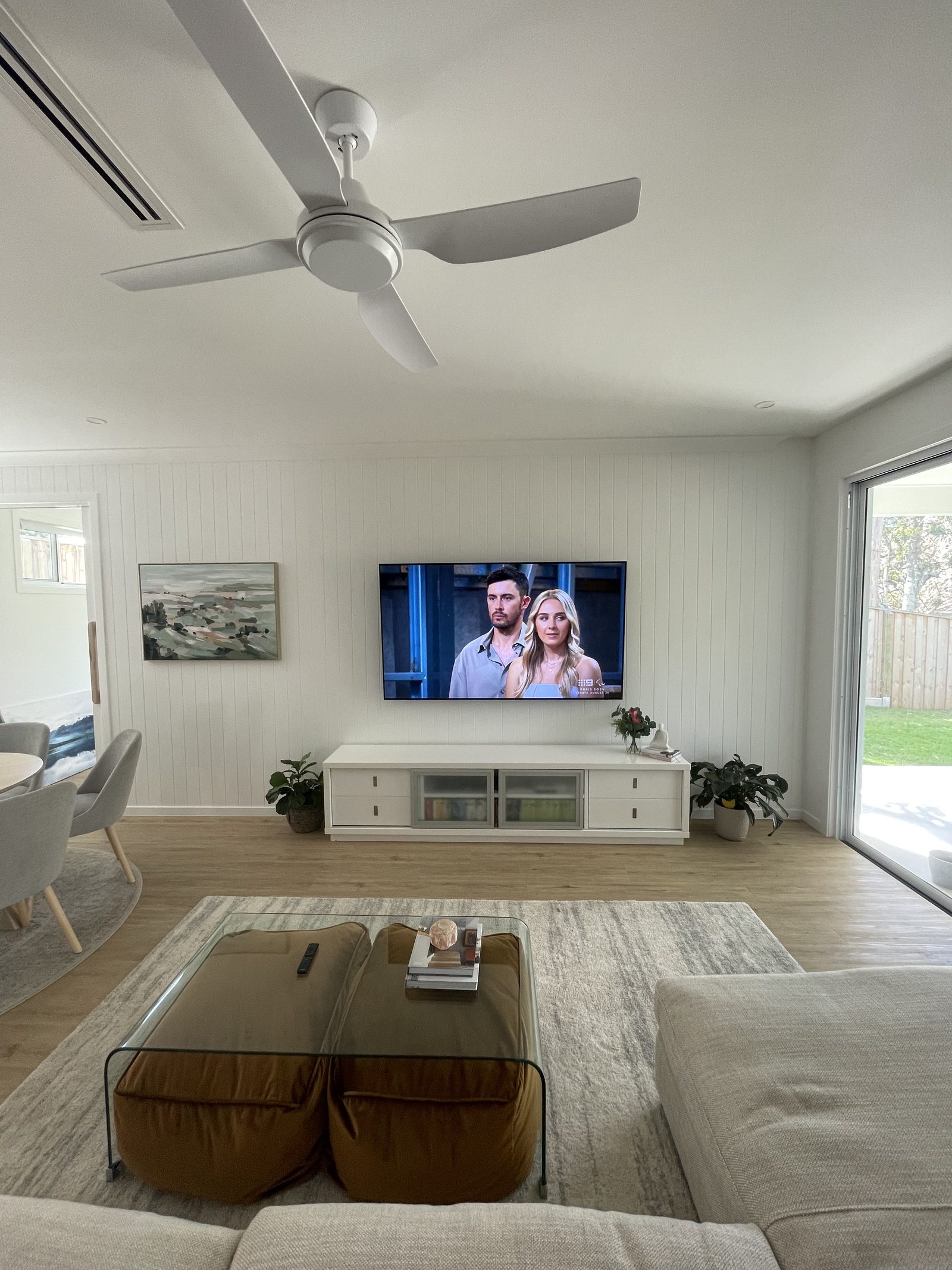 Wall-mounted TV on a white shiplap feature wall with a framed landscape painting hung to the side, set above a long white media console.