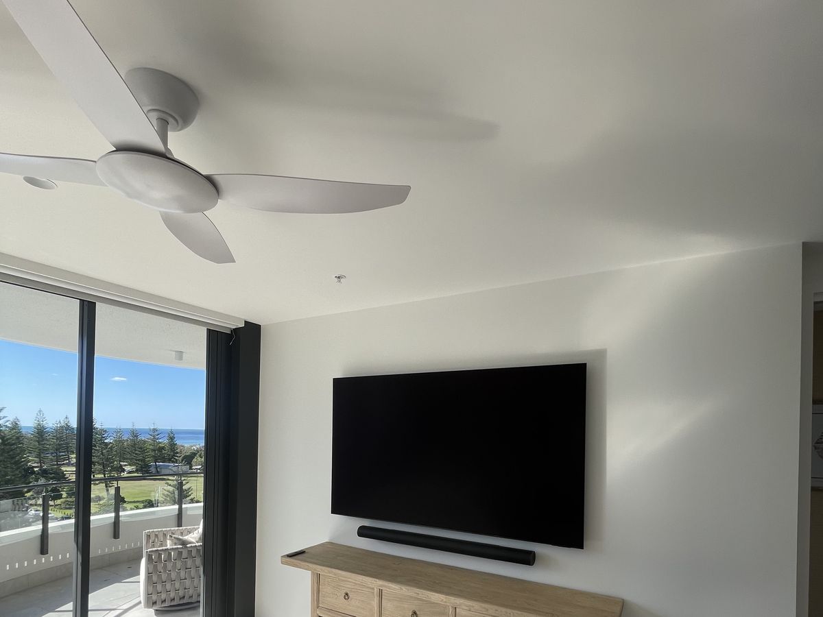 Soundbar mounted level beneath a wall-flush TV in a coastal apartment, ocean view through the balcony beyond. Cliff Hanger Installations.