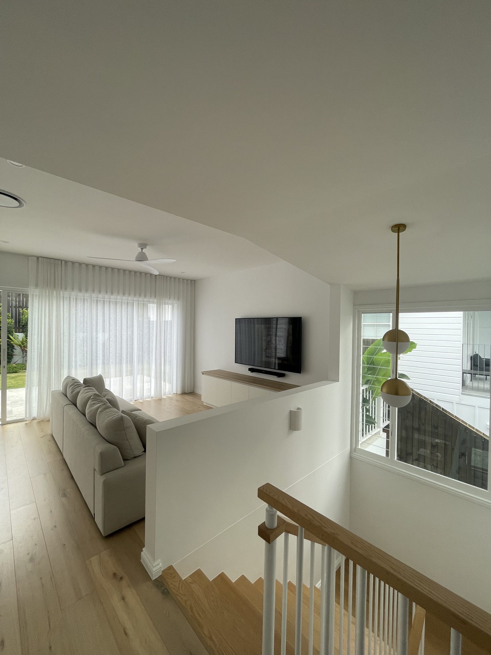 Wall-mounted TV with soundbar in an upstairs living area beside a stair void, with sheer curtains on the windows and a brass pendant light alongside.