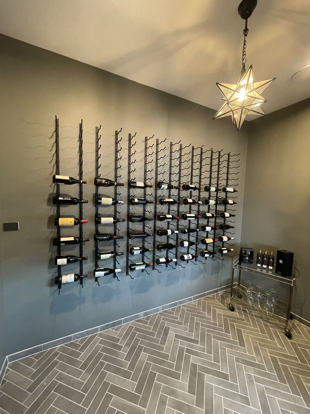 Wall-mounted black metal wine racks holding dozens of bottles beneath a Moravian star pendant in a private wine cellar.